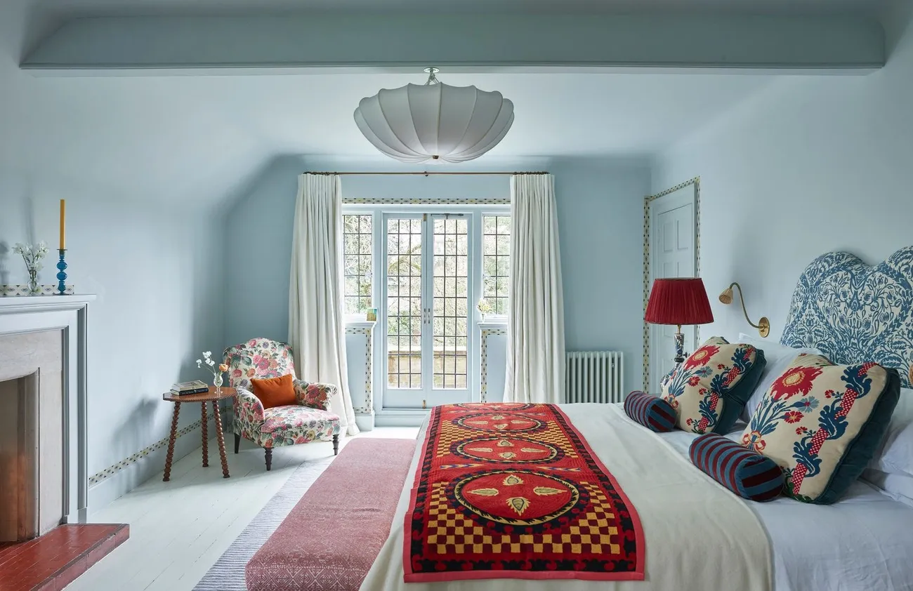 Bright bedroom with floral armchair, patterned pillows, red and orange bed runner, and large window with white curtains.